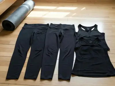 Three pairs of black leggings and two workout tops laid flat side by side on a bedroom floor.