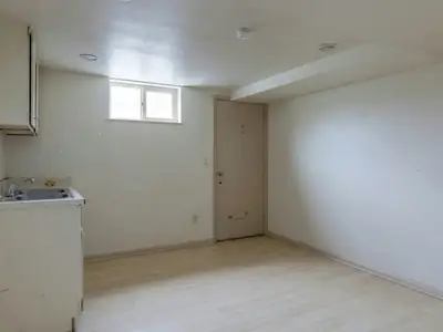 An empty basement suite with a low ceiling, a small window near the ceiling, a compact kitchenette and a separate side entrance door.