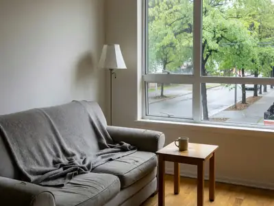 A tidy one-bedroom apartment living room with a loveseat, a floor lamp and a large window looking onto a leafy street.