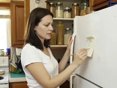 A woman writing a grocery list on a small magnetic notepad stuck to her fridge while checking the pantry shelves.
