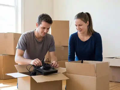 A young couple packing up a modem and cables into a moving box among other packed boxes.
