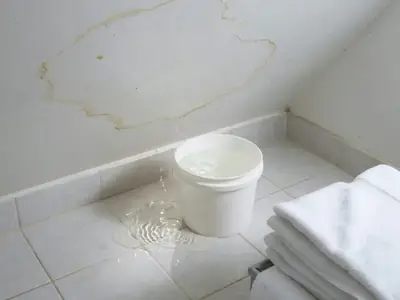 A condo bathroom with water damage on the ceiling and a small puddle on the tile floor below.