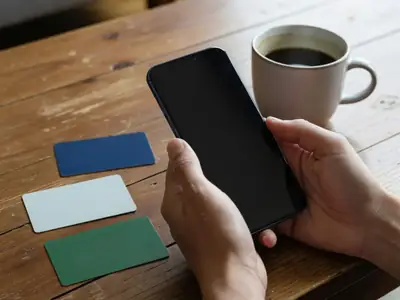 Close-up of hands holding a smartphone with a dark screen over a coffee table, three plain blank bank cards in different colours lying beside it.