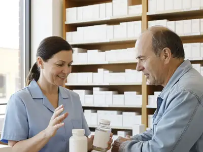 A pharmacist behind a pharmacy counter explaining two plain medicine bottles to a middle-aged customer.