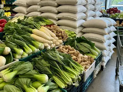 An Asian grocery produce section with bok choy, gai lan, daikon, ginger and stacks of large rice sacks.
