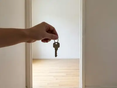 A hand holding out a single key on a plain ring in an empty apartment doorway.
