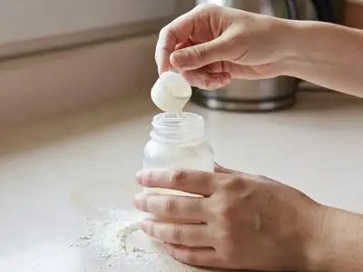 Hands measuring formula powder with a scoop into a baby bottle on a kitchen counter.