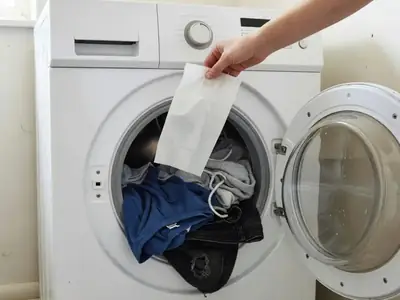 A hand dropping a thin detergent sheet into a washing machine drum full of clothes.