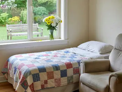 A bright private room in a long-term care home with a bed, a recliner and a window to a garden.