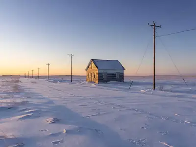 A farmhouse on the open prairie in winter with power lines leading to it.