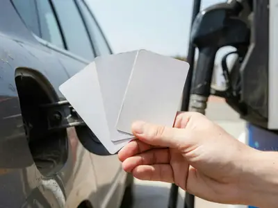 A hand holding three plain blank loyalty cards fanned out beside a car's fuel door.