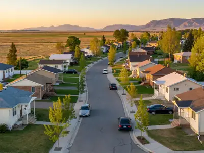 A winding residential street of modest starter homes with mountains or prairie skyline beyond.