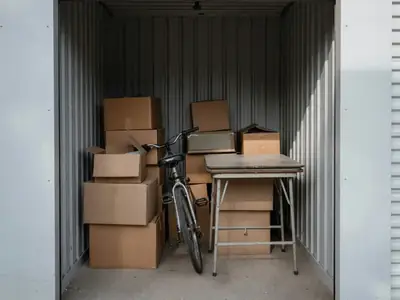 The inside of a small self-storage unit with boxes, a bicycle and a folded table stacked inside.