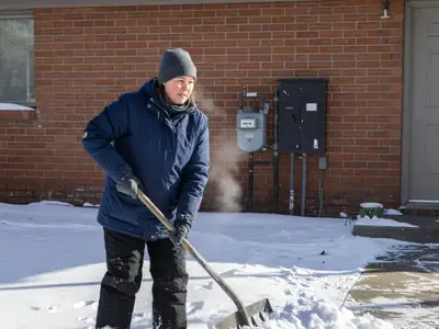 A woman shovelling snow on her front walk with a gas meter and electrical panel on the house wall behind her.