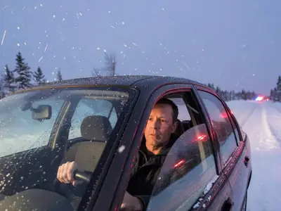 A worried driver sitting behind the wheel of his parked car on a snowy roadside at dusk, hands on the steering wheel, red and blue light reflecting on his face.