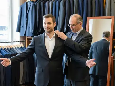 A groom being fitted for a suit jacket by a tailor in a men's clothing shop.